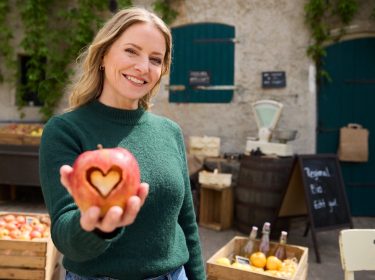 Hauptdarstellerin Diana Staehly steht auf dem Hof und hält lächelnd einen Apfel mit ausgestanztem Herz in der Hand.