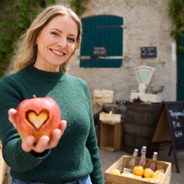 Hauptdarstellerin Diana Staehly steht auf dem Hof und hält lächelnd einen Apfel mit ausgestanztem Herz in der Hand.