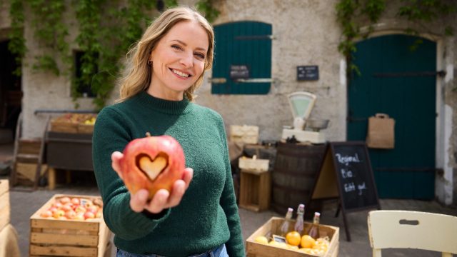 Hauptdarstellerin Diana Staehly steht auf dem Hof und hält lächelnd einen Apfel mit ausgestanztem Herz in der Hand.