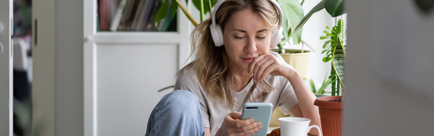 Eine junge Frau sitzt entspannt an einem Tisch im Wohnzimmer, umgeben von Zimmerpflanzen. Sie trägt weiße Kopfhörer, schaut auf ihr Smartphone und hört offenbar konzentriert einen Beitrag. Vor ihr stehen ein Croissant und eine weiße Tasse. Die Szene vermittelt Ruhe und Fokus – passend zum Thema beste ARD-Podcasts.