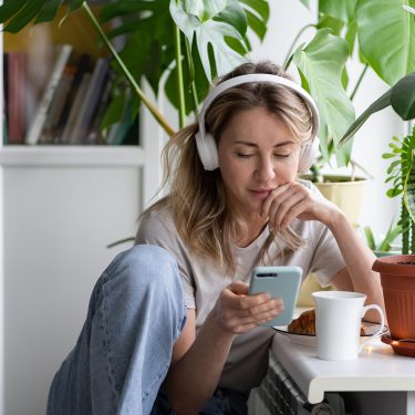 Eine junge Frau sitzt entspannt an einem Tisch im Wohnzimmer, umgeben von Zimmerpflanzen. Sie trägt weiße Kopfhörer, schaut auf ihr Smartphone und hört offenbar konzentriert einen Beitrag. Vor ihr stehen ein Croissant und eine weiße Tasse. Die Szene vermittelt Ruhe und Fokus – passend zum Thema beste ARD-Podcasts.