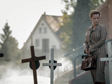 Die Bleikinder bei Netflix: Eine besorgte Frau steht mit einer Handtasche in den Händen an Kindergräbern vor einer Kirche.