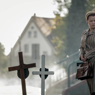 Die Bleikinder bei Netflix: Eine besorgte Frau steht mit einer Handtasche in den Händen an Kindergräbern vor einer Kirche.