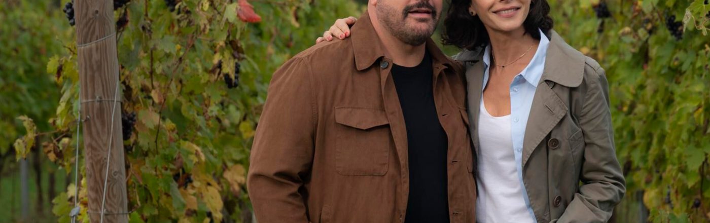 Auf dem Bild zum Film Solo Mio mit Kevin James stehen Matt, gespielt von Kevin James, und Gia, gespielt von Nicole Grimaudo, eng beieinander inmitten eines Weinbergs. Beide wirken gelöst und glücklich – Matt trägt eine braune Jacke, Gia einen beigen Mantel. Die herbstlich gefärbten Reben im Hintergrund erzeugen eine ruhige und romantische Stimmung.