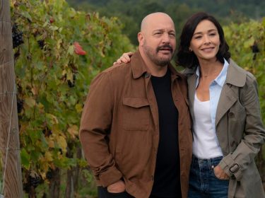 Auf dem Bild zum Film Solo Mio mit Kevin James stehen Matt, gespielt von Kevin James, und Gia, gespielt von Nicole Grimaudo, eng beieinander inmitten eines Weinbergs. Beide wirken gelöst und glücklich – Matt trägt eine braune Jacke, Gia einen beigen Mantel. Die herbstlich gefärbten Reben im Hintergrund erzeugen eine ruhige und romantische Stimmung.