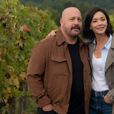 Auf dem Bild zum Film Solo Mio mit Kevin James stehen Matt, gespielt von Kevin James, und Gia, gespielt von Nicole Grimaudo, eng beieinander inmitten eines Weinbergs. Beide wirken gelöst und glücklich – Matt trägt eine braune Jacke, Gia einen beigen Mantel. Die herbstlich gefärbten Reben im Hintergrund erzeugen eine ruhige und romantische Stimmung.