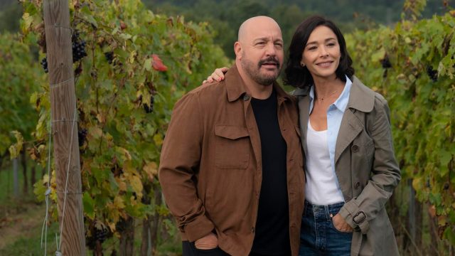 Auf dem Bild zum Film Solo Mio mit Kevin James stehen Matt, gespielt von Kevin James, und Gia, gespielt von Nicole Grimaudo, eng beieinander inmitten eines Weinbergs. Beide wirken gelöst und glücklich – Matt trägt eine braune Jacke, Gia einen beigen Mantel. Die herbstlich gefärbten Reben im Hintergrund erzeugen eine ruhige und romantische Stimmung.