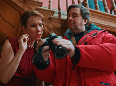 In den Streaming-Highlights im Februar 2026 ist eine Szene aus „Fabian und Die mörderische Hochzeit“ zu sehen. Zu sehen ist Bastian Pastewka, der in einer roten Jacke eine Kamera prüft, während eine Frau neben ihm gespannt hinschaut.