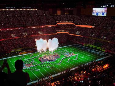 Auf dem Bild in den GigaTV-Highlights im Februar 2026 siehst Du eine Szene vom Super Bowl. In einem riesigen Stadion sind tausende Zuschauer zu sehen, während auf dem Spielfeld eine spektakuläre Halbzeitshow mit Rauch, Licht und Choreografien stattfindet. Die Stimmung wirkt energiegeladen, monumental und voller sportlicher und musikalischer Highlights.
