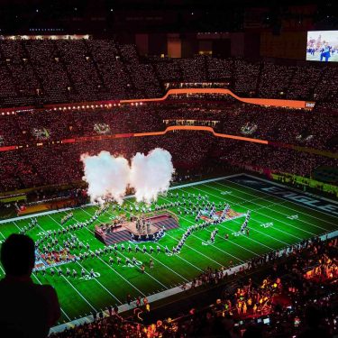 Auf dem Bild in den GigaTV-Highlights im Februar 2026 siehst Du eine Szene vom Super Bowl. In einem riesigen Stadion sind tausende Zuschauer zu sehen, während auf dem Spielfeld eine spektakuläre Halbzeitshow mit Rauch, Licht und Choreografien stattfindet. Die Stimmung wirkt energiegeladen, monumental und voller sportlicher und musikalischer Highlights.