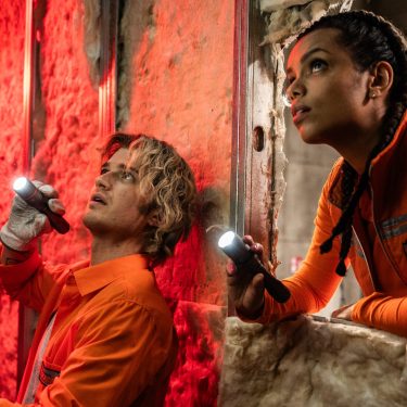 Auf dem Bild zur Cold Storage Filmkritik sind Naomi Williams, gespielt von Georgina Campbell, und Travis „Teacake“ Meacham, dargestellt von Joe Keery, in einer spannungsgeladenen Filmszene zu sehen. Beide tragen orange Arbeitskleidung und leuchten mit Taschenlampen in einen dunklen, industriellen Raum. Die rote Beleuchtung und ihre konzentrierten Blicke erzeugen eine bedrohliche, nervöse Atmosphäre.