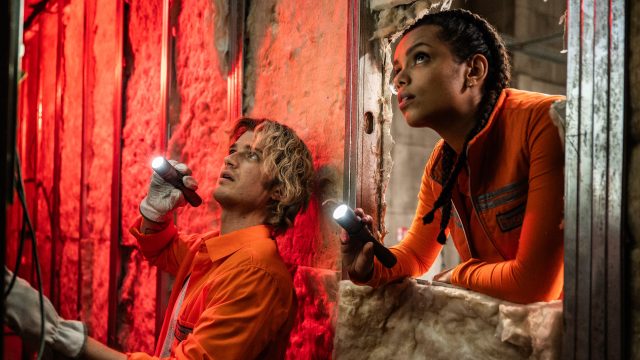 Auf dem Bild zur Cold Storage Filmkritik sind Naomi Williams, gespielt von Georgina Campbell, und Travis „Teacake“ Meacham, dargestellt von Joe Keery, in einer spannungsgeladenen Filmszene zu sehen. Beide tragen orange Arbeitskleidung und leuchten mit Taschenlampen in einen dunklen, industriellen Raum. Die rote Beleuchtung und ihre konzentrierten Blicke erzeugen eine bedrohliche, nervöse Atmosphäre.