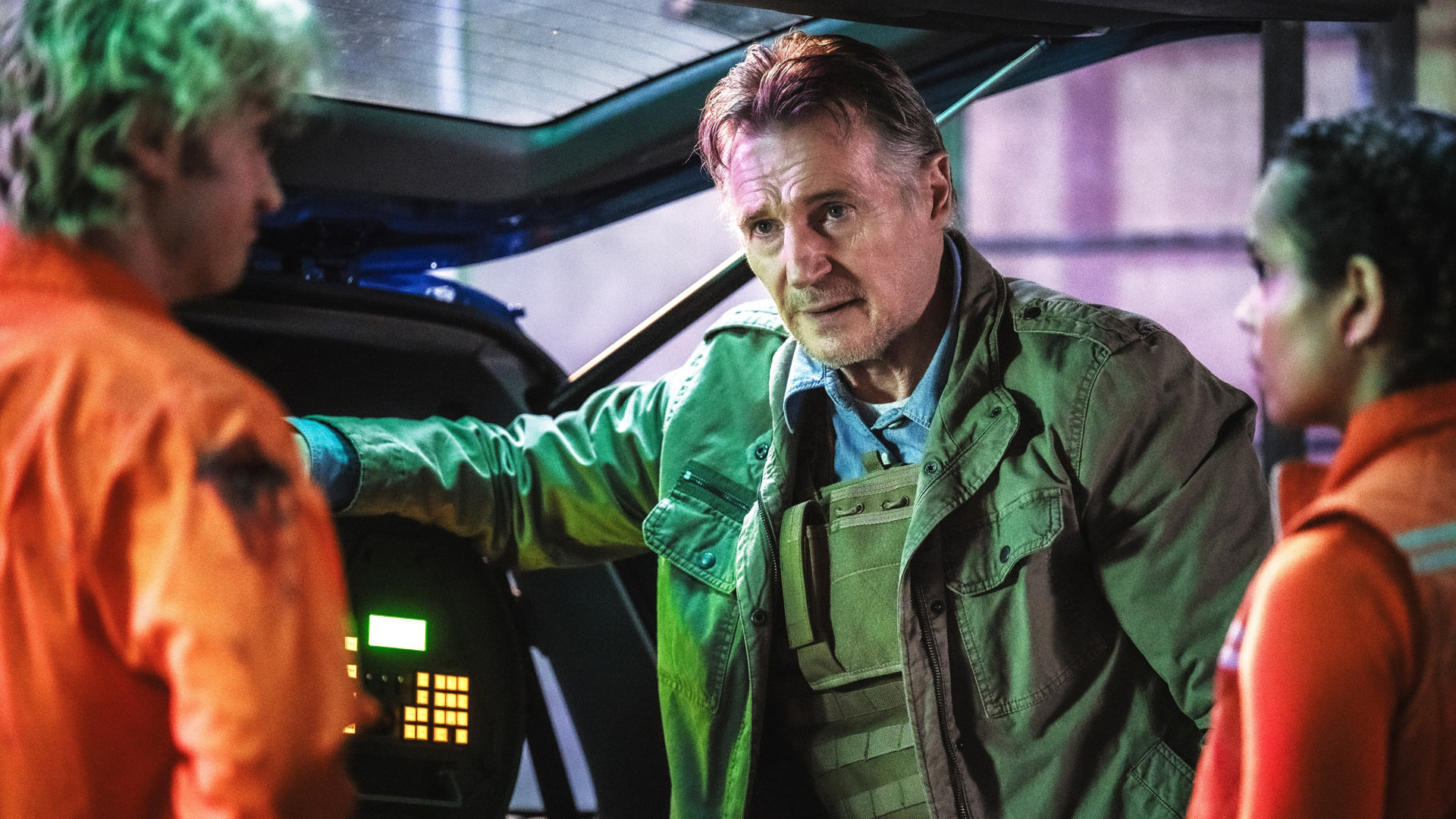 Auf dem Bild zur Cold Storage Filmkritik ist ein Bioterror-Agent zu sehen, gespielt von Liam Neeson, der ernst und kontrolliert vor zwei Personen in orangefarbener Schutzkleidung steht. In einer düsteren, technisch anmutenden Umgebung lehnt er an einem geöffneten Fahrzeug oder Gerät. Die grünliche Beleuchtung und seine angespannte Mimik vermitteln eine bedrohliche, konfliktreiche Stimmung.