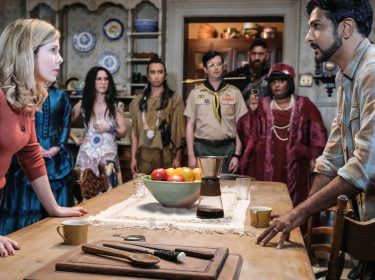 Samantha (Rose McIver) und ihr Partner Jay (Utkarsh Ambudkar) stehen mit ihren eigentümlichen Geister-Mitbewohner:innen um einen Tisch herum versammelt.