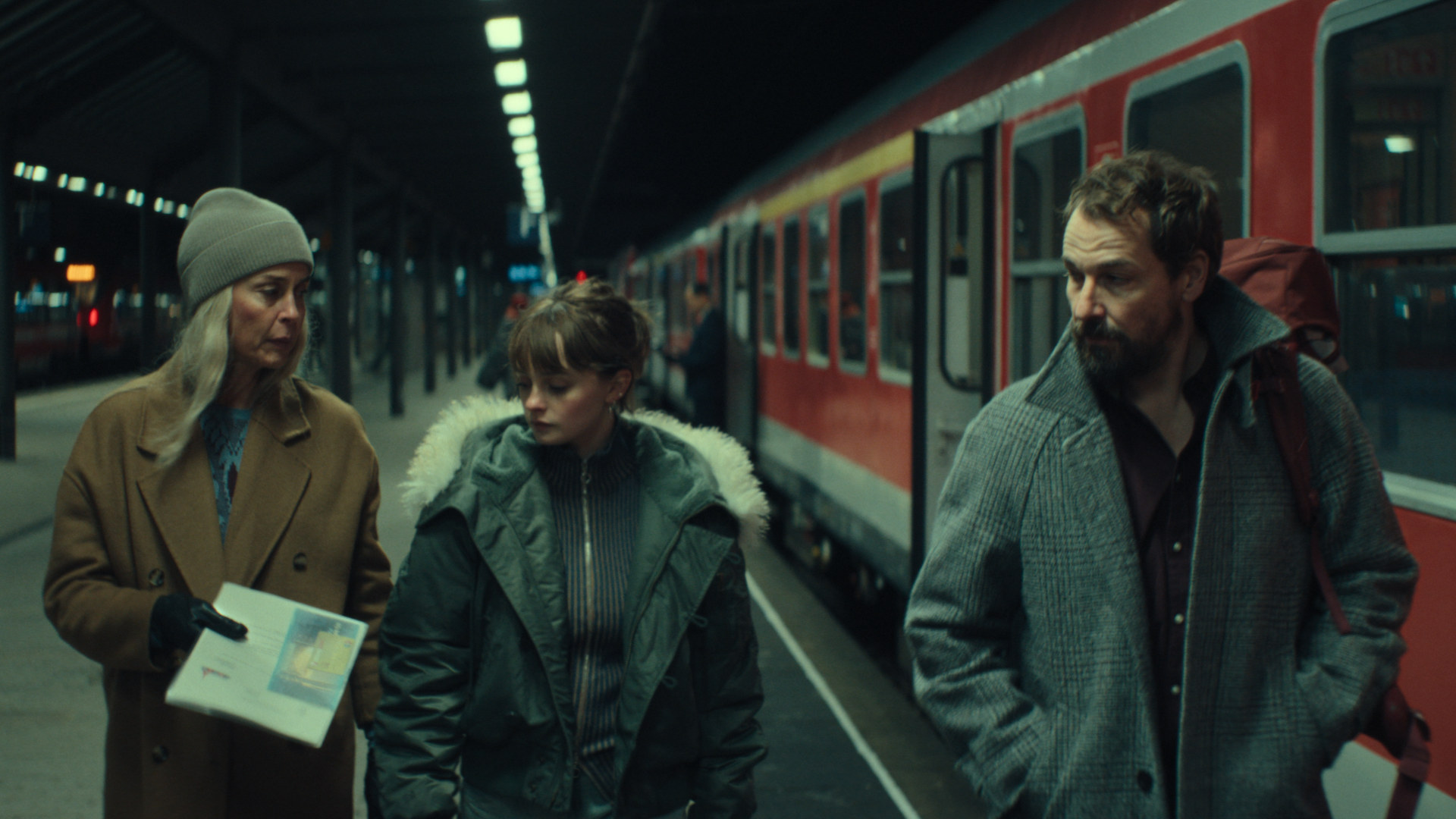 Meret und Simon gehen mit ihrer Tochter über einen Bahnhof und unterhalten sich dabe.