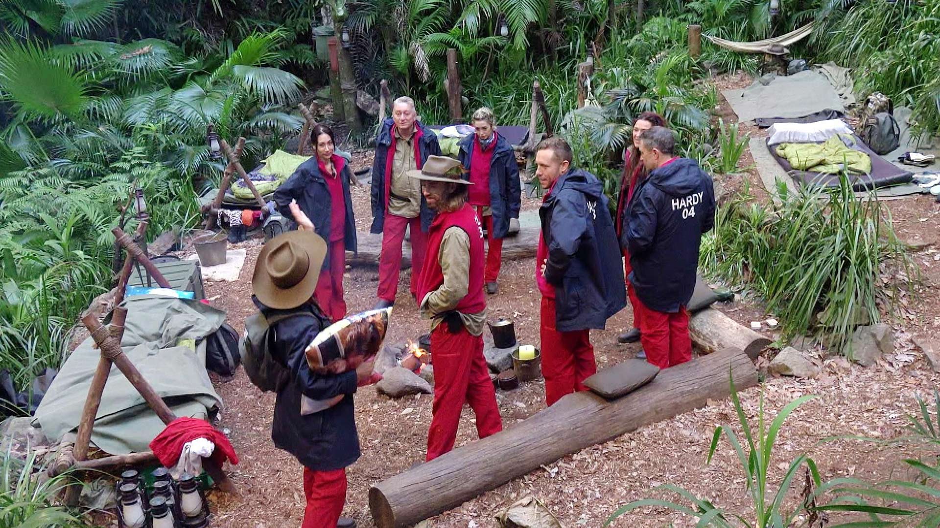 Auf dem Bild zum Dschungelcamp 2026 sind mehrere Kandidatinnen und Kandidaten im australischen Dschungelcamp zu sehen. Sie stehen in roter Campkleidung im Kreis zusammen, umgeben von Schlafplätzen, Lagerfeuer und dichter Vegetation. Die Stimmung wirkt angespannt und konzentriert, als würde eine wichtige Besprechung oder ein Moment nach einer Prüfung stattfinden.