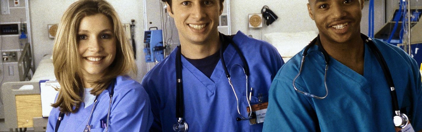 Auf dem Bild zu Scrubs beste Folgen sind drei Hauptcharaktere der Serie Scrubs in einem Krankenhaus zu sehen. Links steht Elliot, gespielt von Sarah Chalke, freundlich lächelnd in blauer OP-Kleidung. In der Mitte J.D., dargestellt von Zac Braff, locker und optimistisch. Rechts steht Turk, gespielt von Donald Faison, selbstbewusst und gut gelaunt.