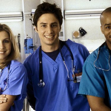 Auf dem Bild zu Scrubs beste Folgen sind drei Hauptcharaktere der Serie Scrubs in einem Krankenhaus zu sehen. Links steht Elliot, gespielt von Sarah Chalke, freundlich lächelnd in blauer OP-Kleidung. In der Mitte J.D., dargestellt von Zac Braff, locker und optimistisch. Rechts steht Turk, gespielt von Donald Faison, selbstbewusst und gut gelaunt.