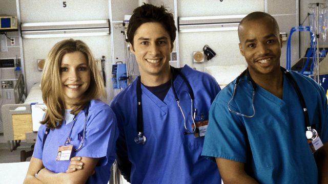 Auf dem Bild zu Scrubs beste Folgen sind drei Hauptcharaktere der Serie Scrubs in einem Krankenhaus zu sehen. Links steht Elliot, gespielt von Sarah Chalke, freundlich lächelnd in blauer OP-Kleidung. In der Mitte J.D., dargestellt von Zac Braff, locker und optimistisch. Rechts steht Turk, gespielt von Donald Faison, selbstbewusst und gut gelaunt.
