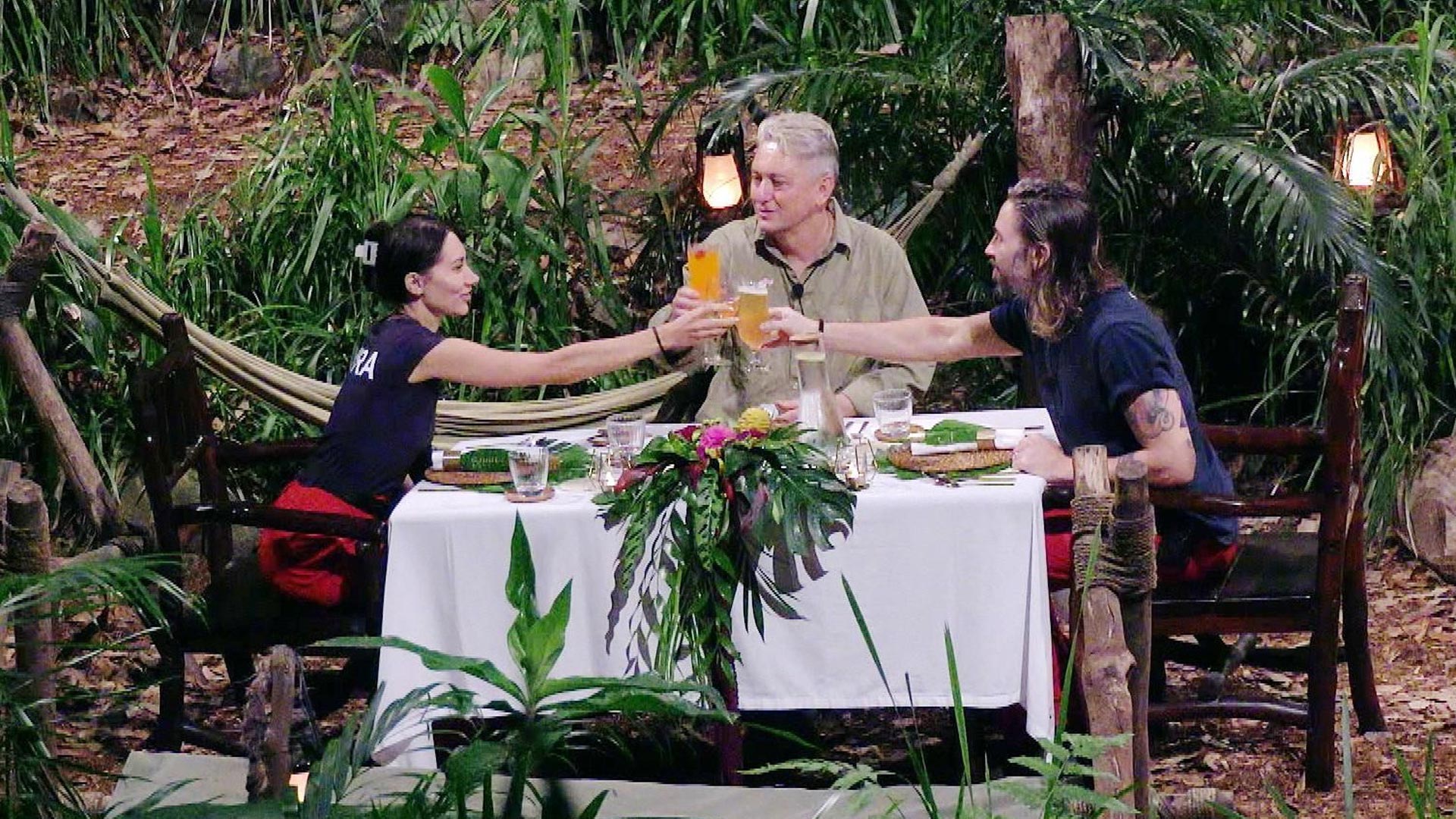 Auf dem Bild zum Dschungelcamp 2026 ist das Finale Dinner mit Samira, Hubert und Gil zu sehen. Die drei sitzen an einem festlich gedeckten Tisch mitten im Dschungel und stoßen mit Getränken an. Umgeben von Pflanzen wirkt die Szene harmonisch, erleichtert und feierlich nach den intensiven Tagen im Camp.