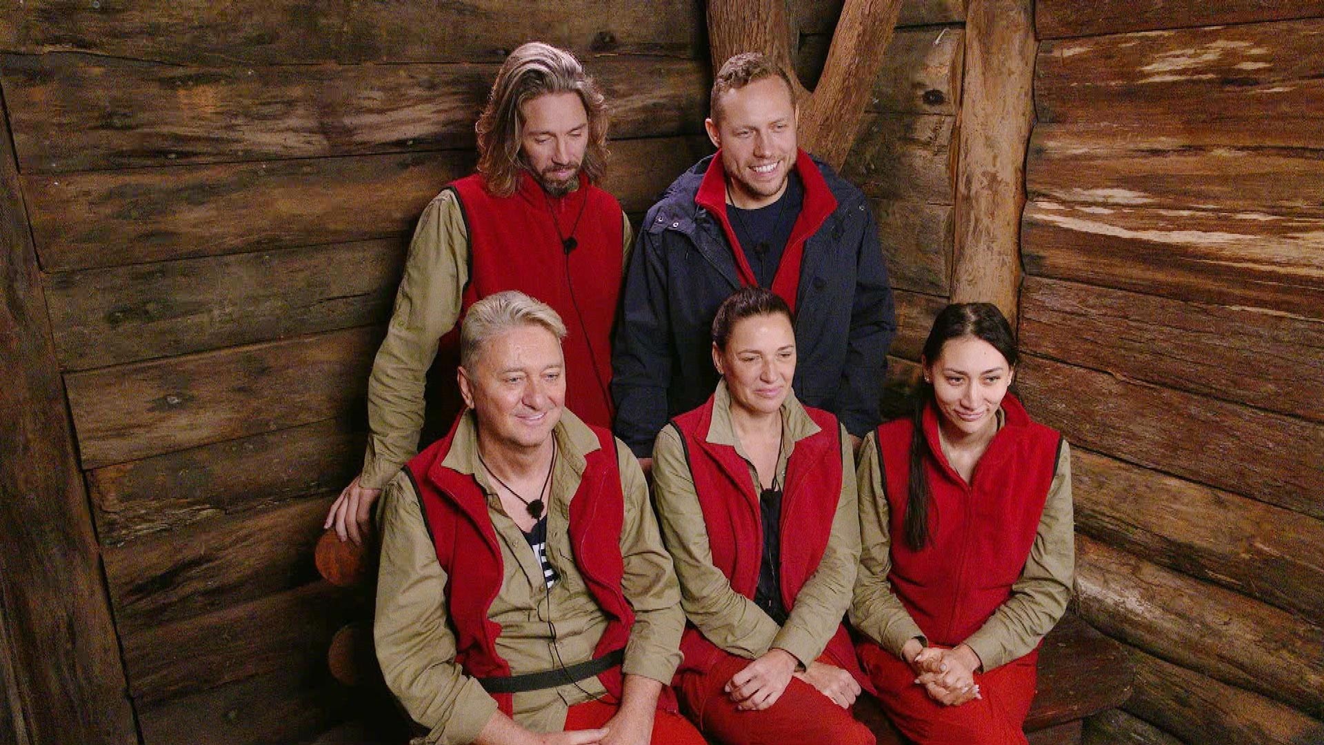 Auf dem Bild zum Dschungelcamp 2026 sind die Halbfinalisten Gil, Patrick, Hubert, Simone und Samira in einer gemeinsamen Szene zu sehen. Die Gruppe sitzt eng beieinander in einer hölzernen Dschungelprüfungskulisse und trägt rote Westen. Die Stimmung wirkt ruhig, angespannt und erwartungsvoll vor der nächsten Entscheidung.