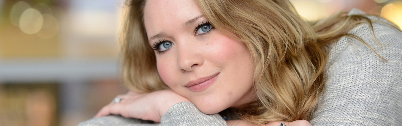 Auf dem Bild zur Crescent City-Reihenfolge ist die Autorin Sarah J. Maas in einem ruhigen Porträt zu sehen. Sie lehnt entspannt an einer Brüstung, trägt einen hellen Strickpullover und einen auffälligen Ring. Ihr freundlicher Blick und das weiche Bokeh im Hintergrund erzeugen eine warme, inspirierende Stimmung, die Kreativität und literarische Nähe vermittelt.