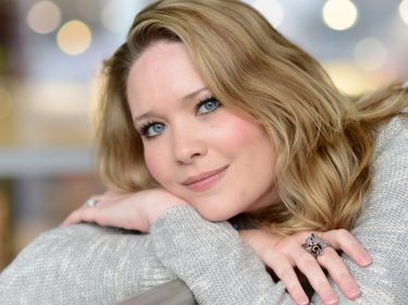 Auf dem Bild zur Crescent City-Reihenfolge ist die Autorin Sarah J. Maas in einem ruhigen Porträt zu sehen. Sie lehnt entspannt an einer Brüstung, trägt einen hellen Strickpullover und einen auffälligen Ring. Ihr freundlicher Blick und das weiche Bokeh im Hintergrund erzeugen eine warme, inspirierende Stimmung, die Kreativität und literarische Nähe vermittelt.