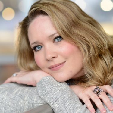 Auf dem Bild zur Crescent City-Reihenfolge ist die Autorin Sarah J. Maas in einem ruhigen Porträt zu sehen. Sie lehnt entspannt an einer Brüstung, trägt einen hellen Strickpullover und einen auffälligen Ring. Ihr freundlicher Blick und das weiche Bokeh im Hintergrund erzeugen eine warme, inspirierende Stimmung, die Kreativität und literarische Nähe vermittelt.