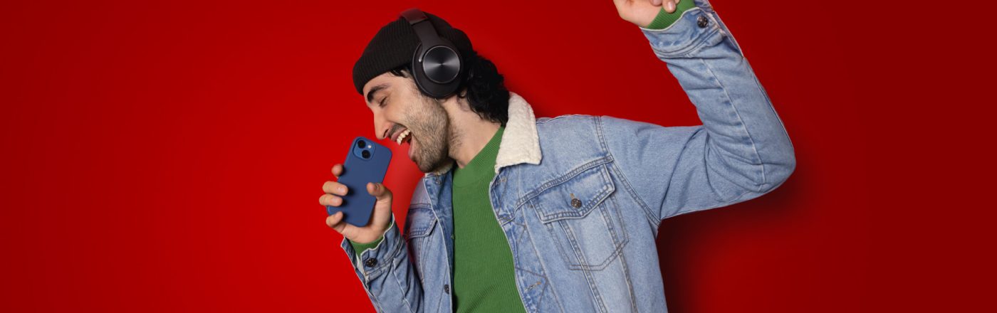 Auf dem Bild in der Musik-Streamingdienste-Übersicht ist ein junger Mann vor rotem Hintergrund zu sehen, der mit geschlossenen Augen begeistert in ein Smartphone singt. Er trägt schwarze Over-Ear-Kopfhörer, eine schwarze Mütze, ein grünes Shirt und eine Jeansjacke. Seine erhobene Faust unterstreicht die ausgelassene, energiegeladene Stimmung beim Musikhören.