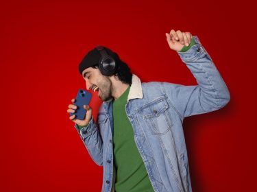 Auf dem Bild in der Musik-Streamingdienste-Übersicht ist ein junger Mann vor rotem Hintergrund zu sehen, der mit geschlossenen Augen begeistert in ein Smartphone singt. Er trägt schwarze Over-Ear-Kopfhörer, eine schwarze Mütze, ein grünes Shirt und eine Jeansjacke. Seine erhobene Faust unterstreicht die ausgelassene, energiegeladene Stimmung beim Musikhören.