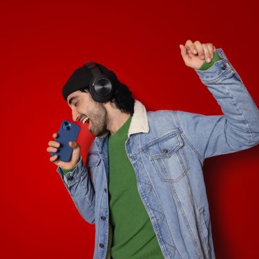 Auf dem Bild in der Musik-Streamingdienste-Übersicht ist ein junger Mann vor rotem Hintergrund zu sehen, der mit geschlossenen Augen begeistert in ein Smartphone singt. Er trägt schwarze Over-Ear-Kopfhörer, eine schwarze Mütze, ein grünes Shirt und eine Jeansjacke. Seine erhobene Faust unterstreicht die ausgelassene, energiegeladene Stimmung beim Musikhören.