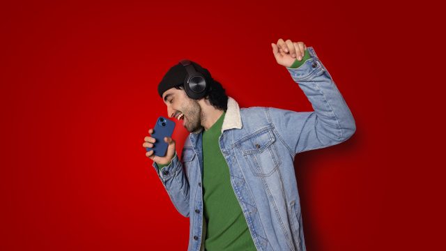 Auf dem Bild in der Musik-Streamingdienste-Übersicht ist ein junger Mann vor rotem Hintergrund zu sehen, der mit geschlossenen Augen begeistert in ein Smartphone singt. Er trägt schwarze Over-Ear-Kopfhörer, eine schwarze Mütze, ein grünes Shirt und eine Jeansjacke. Seine erhobene Faust unterstreicht die ausgelassene, energiegeladene Stimmung beim Musikhören.