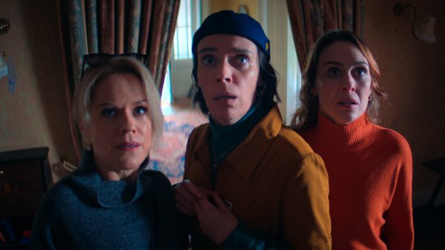 Szenenbild aus Hot To Get To Heaven From Belfast: Drei Frauen stehen mit schockiertem Blick in einer dramatischen Szene in einem Wohnzimmer vor einem Vorhang.