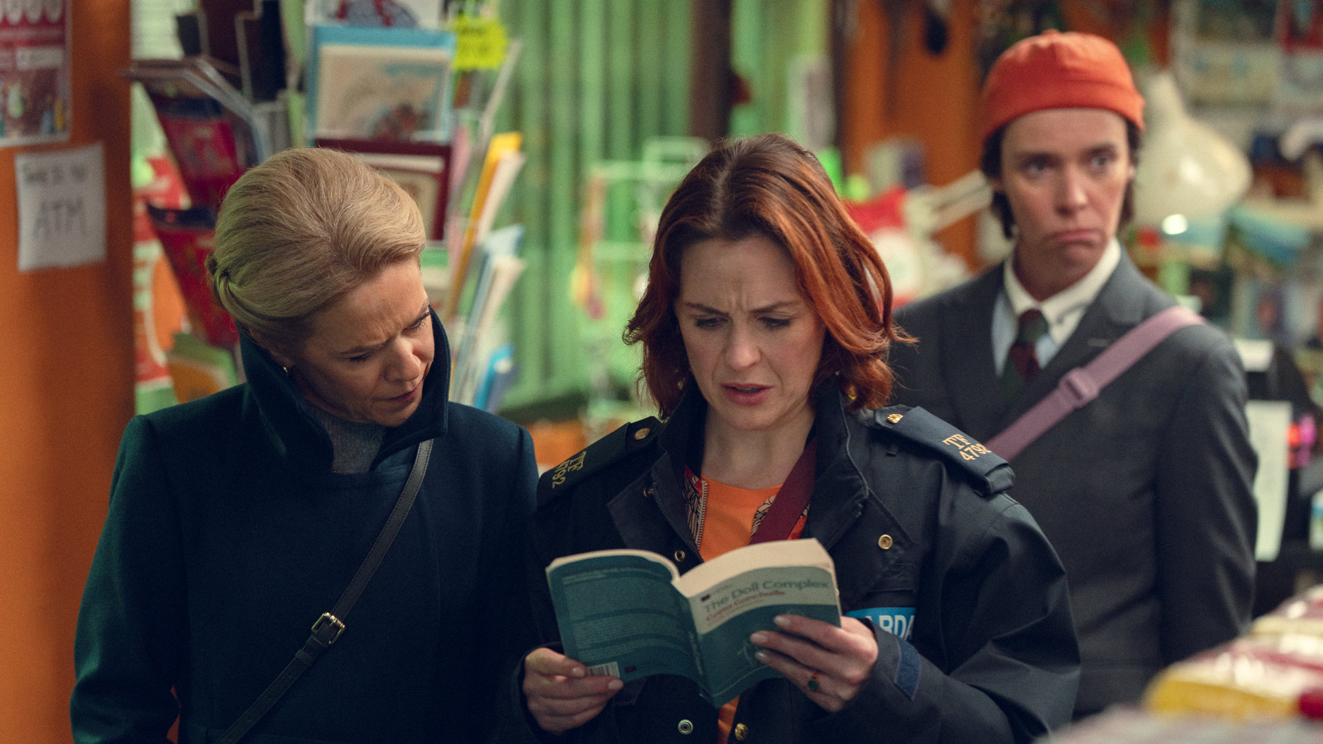 Szenenbild aus Hot To Get To Heaven From Belfast: Zwei Frauen blicken konzentriert in ein Buch mit der Aufschrift „The Doll Complex“ während eine Dritte mit nachdenklicher Miene im Hintergrund steht.