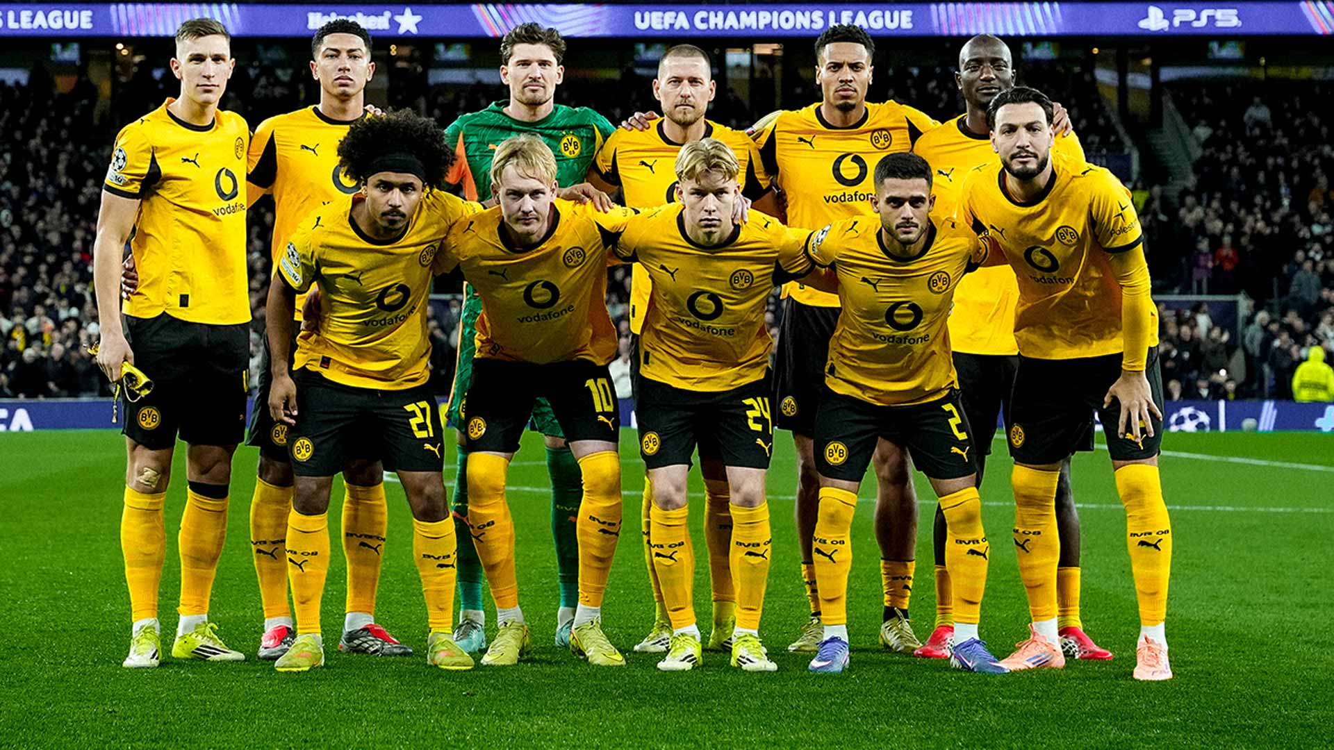 Im Bild vom BVB-Mediathek-Artikel ist die Mannschaft von Borussia Dortmund vor einem UEFA-Champions-League-Spiel zu sehen. Elf Spieler stehen in zwei Reihen auf dem Spielfeld, tragen gelbe Trikots mit schwarzer Hose und gelben Stutzen. Im Hintergrund sind das Stadion, Zuschauer sowie ein Banner mit der Aufschrift „UEFA Champions League“ erkennbar.