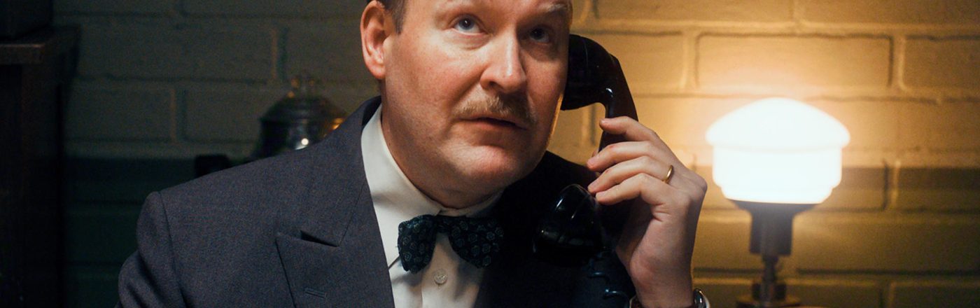 Henrik Dorsin in Die Schwedische Verbindung Ein Mann mit Schnurrbart und fliehender Stirn hält einen Telefonhörer am Ohr.
