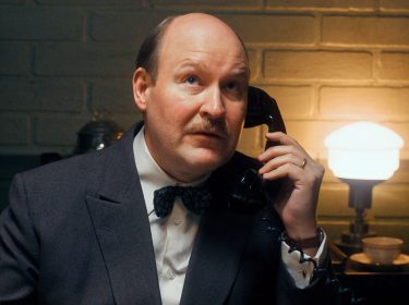 Henrik Dorsin in Die Schwedische Verbindung Ein Mann mit Schnurrbart und fliehender Stirn hält einen Telefonhörer am Ohr.