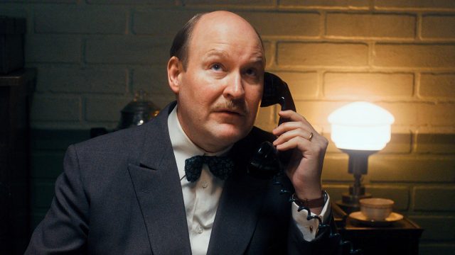 Henrik Dorsin in Die Schwedische Verbindung Ein Mann mit Schnurrbart und fliehender Stirn hält einen Telefonhörer am Ohr.