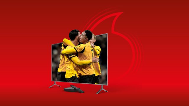 Auf dem Werbebild zur BVB-Mediathek ist ein Fernseher vor rotem Hintergrund abgebildet, auf dessen Bildschirm mehrere BVB-Spieler in gelben Trikots zu sehen sind, die sich umarmen. Unten ist eine Fernbedienung vor dem Fernseher platziert.